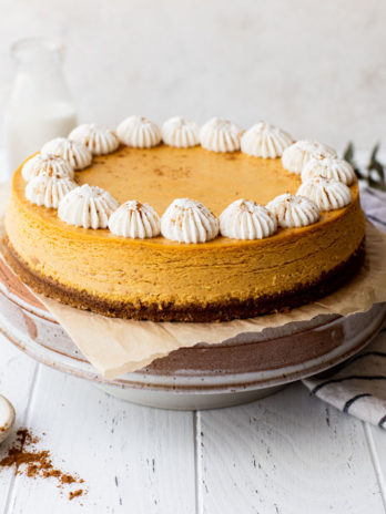 A pumpkin cheesecake on top of a cake stand with milk and cinnamon around it.